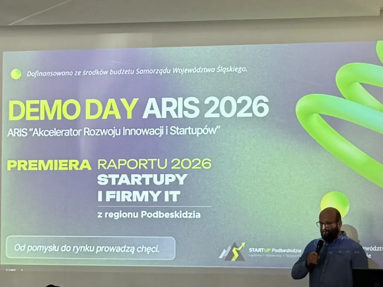 Slajd otwierający Demo Day ARIS 2026 – Akcelerator Rozwoju Innowacji i Startupów, z zapowiedzią premiery raportu 2026 „Startupy i firmy IT z regionu Podbeskidzia”; obok sceny prowadzący z mikrofonem